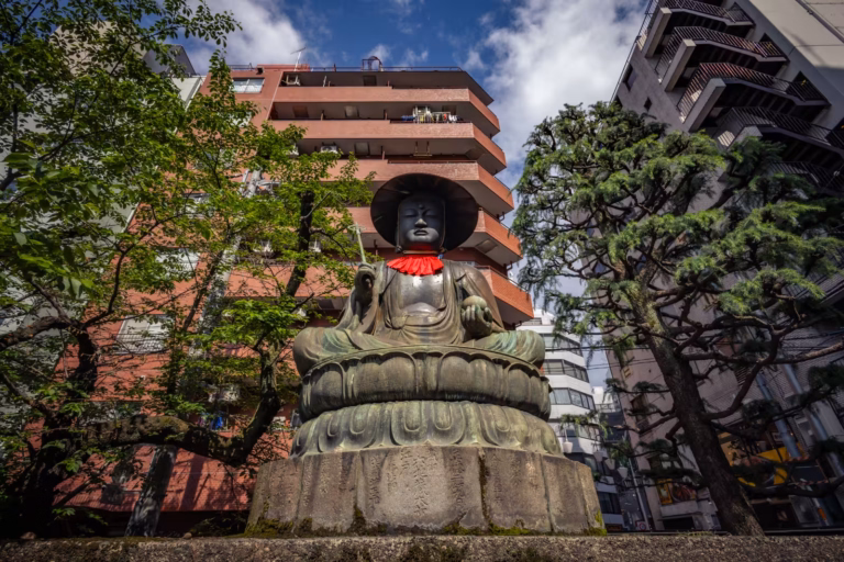 Low-angle view of a seated bronze Jizo statue with a black hat and red bib, framed by urban buildings and leafy trees.