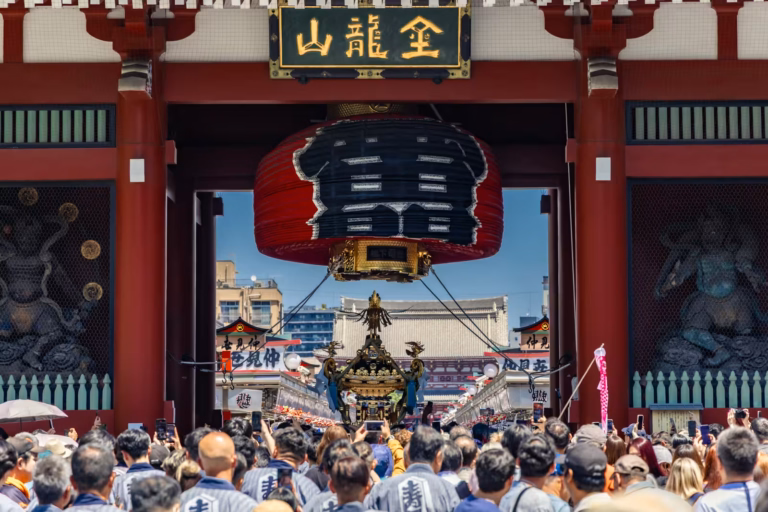 Crowds in happi coats carry a golden mikoshi under the Kaminarimon gate's giant red lantern during a festival.