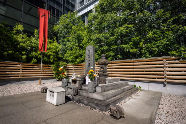 Full view of the Masakado-kubizuka shrine in Tokyo, showing the stone monument, red banner, and protective frog statues.