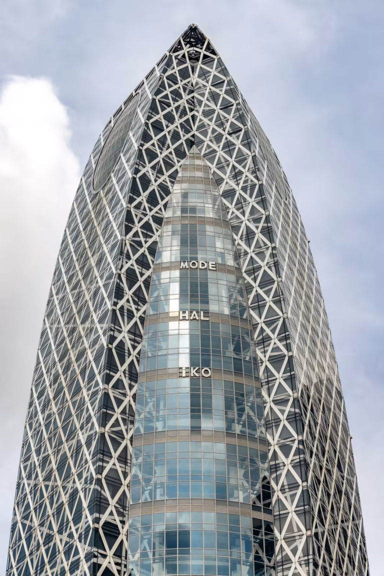Low-angle view of the Mode Gakuen Cocoon Tower's white lattice and curved glass facade against a soft, cloudy sky.
