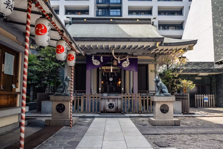 Kitsune statues guard the entrance to Miyasmasumitake Shrine, set against a backdrop of modern urban apartments.