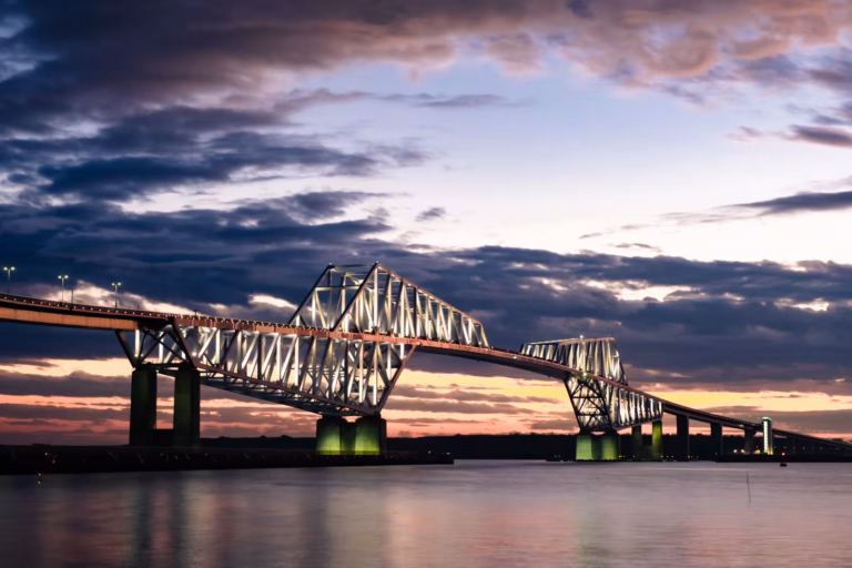 The illuminated Tokyo Gate Bridge glows against a purple and orange sunset sky over calm, reflective water.