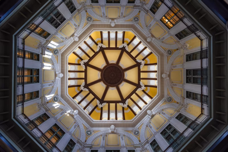 Symmetrical upward view of a yellow octagonal dome with brown ribs, white eagle statues, and architectural tiers.
