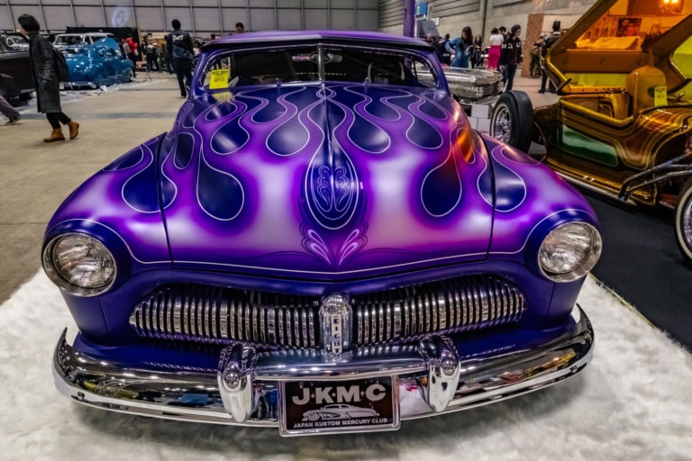 Front view of a custom purple Mercury with pinstriped flames and a chrome grille at an indoor car show.