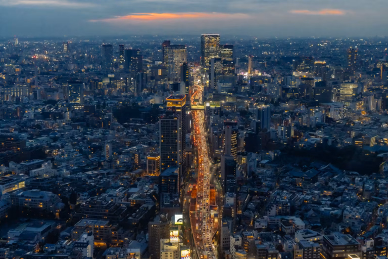 Aerial night view of a dense city with a central glowing highway of traffic under a faint orange sunset horizon.
