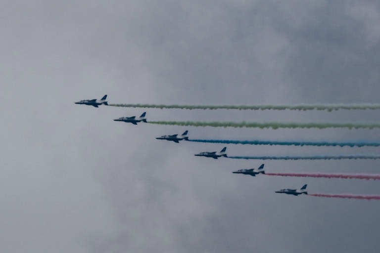 Six Blue Impulse jets in formation, trailing green, blue, and red smoke against an overcast sky.