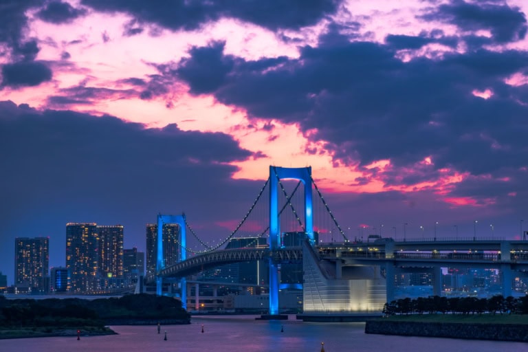 Tokyo’s Rainbow Bridge glows blue against a dramatic pink and purple twilight sky with city lights.