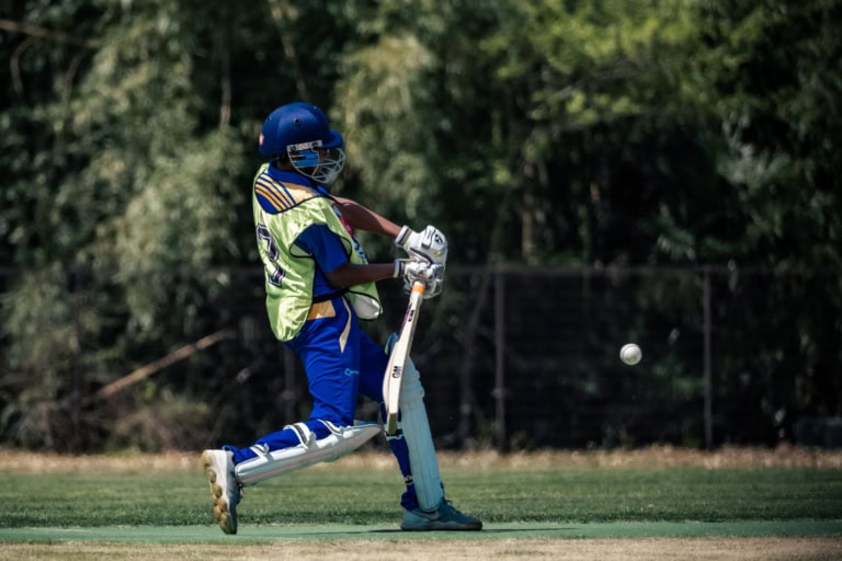 Action shot of a cricket batsman in blue gear and a yellow vest swinging at an approaching ball on a grassy field.