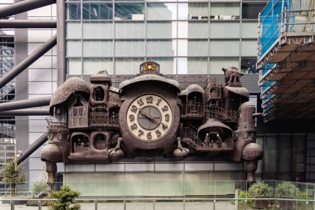 Massive copper steampunk clock with a large face and mechanical compartments on the side of a modern glass skyscraper.