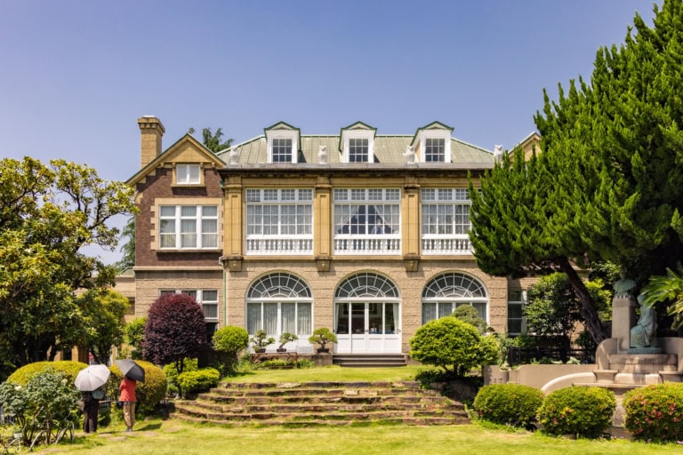 Hatoyama Hall, a grand stone mansion with arched windows, sits behind a green garden lawn with stone steps and trees.