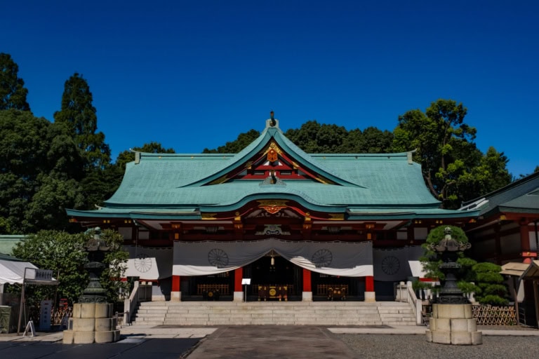 The Hie Shrine main hall features a wide teal roof, red pillars, and white drapes against a clear blue sky.