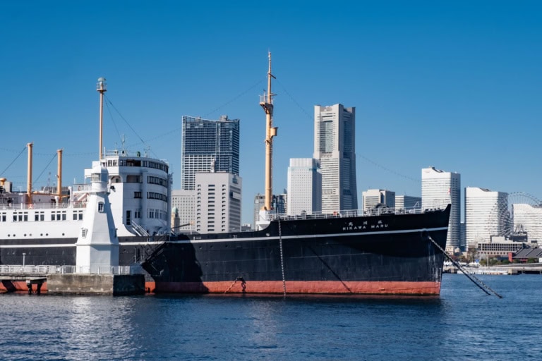 The Hikawa Maru docked in Yokohama harbor, its black hull contrasted against the city skyline under a clear blue sky.