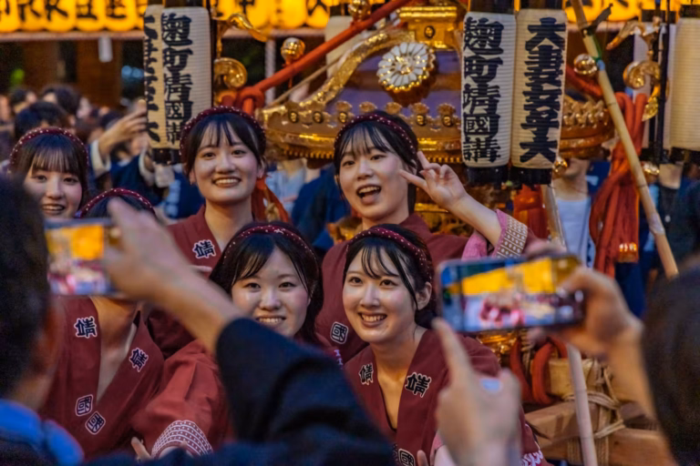 Five women in red festival attire pose for photos in front of a golden mikoshi during Tokyo’s Mitama Matsuri.