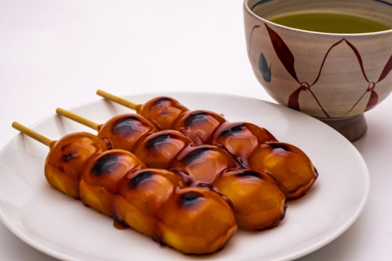 Skewers of glossy, charred mitarashi dango on a white plate beside a traditional ceramic cup of green tea.