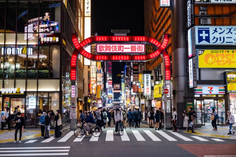 People cross a busy street beneath the iconic red neon Kabukicho gate in Shinjuku at night.