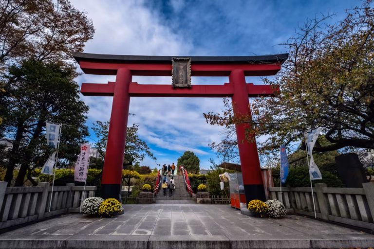 A large red torii gate marks the entrance to a Japanese shrine, with visitors ascending stairs under a bright sky.