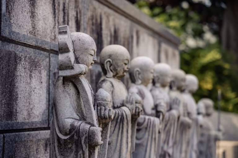 Row of stone Jizo statues standing in prayer against a grey wall at a Japanese temple.