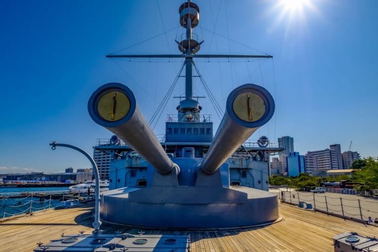 Large twin gun barrels on the wooden deck of the memorial warship Mikasa under a bright sunny sky.