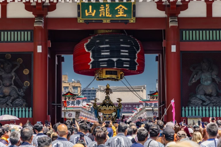 Crowds in happi coats carry a golden mikoshi under the Kaminarimon gate's giant red lantern during a festival.