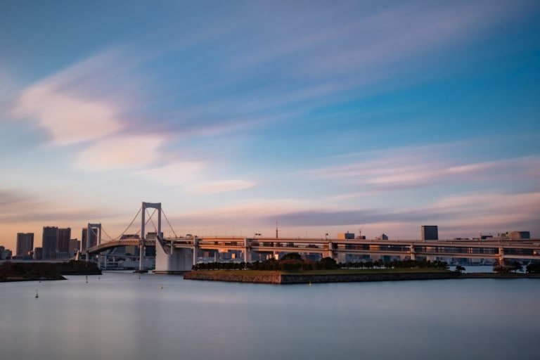 Long exposure of the Rainbow Bridge at sunset with silky water and a pastel sky over the Tokyo city skyline.