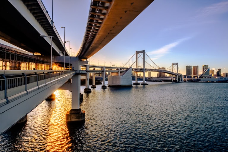 Golden sunset light reflects on Tokyo Bay, framing the Rainbow Bridge beneath layered highway architecture.