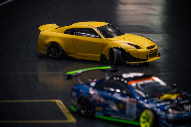 Yellow Nissan GT-R RC drift car sliding on a dark indoor track with a blurred blue racer in the foreground.