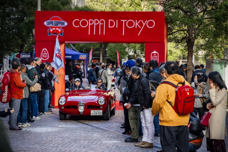 A red vintage Alfa Romeo passes through the Coppa di Tokyo banner as spectators watch on a city street.