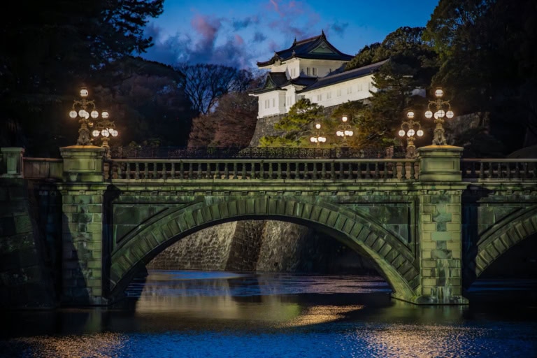 Glowing stone bridge and white turret shine during the Imperial Palace winter illumination, reflecting in the dark water.
