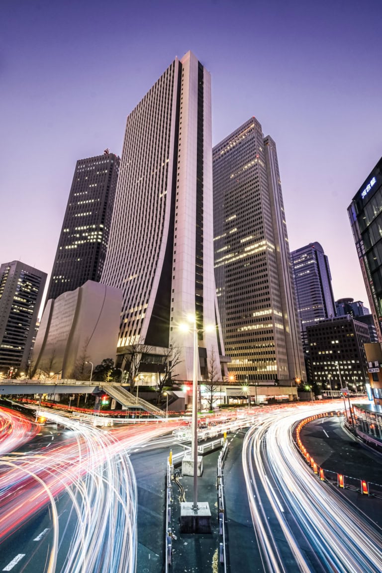 Long exposure light trails streak through a busy Shinjuku intersection below the iconic Sompo Japan building at night.
