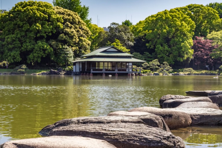Stepping stones lead toward a traditional pavilion across a tranquil pond in Kiyosumi Gardens, framed by lush trees.