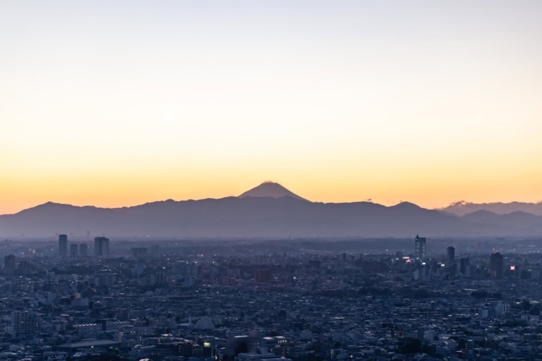Mount Fuji silhouetted against a soft orange sky over a vast, hazy cityscape at twilight.
