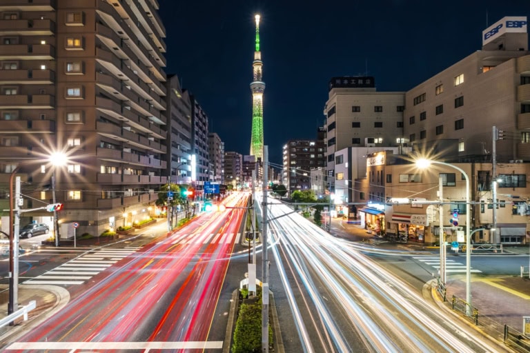 Night long exposure of Tokyo Skytree with light trails like laser beams streaking through a busy urban intersection.