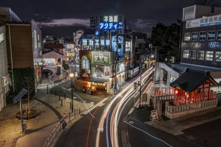 Night long exposure of a city street with light trails passing the Uonami sakaba tavern and a small shrine.