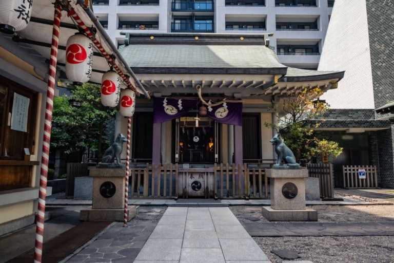 Kitsune statues guard the entrance to Miyasmasumitake Shrine, set against a backdrop of modern urban apartments.