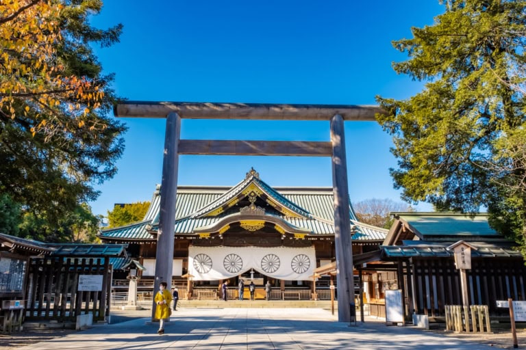 A massive wooden torii gate frames the traditional main hall of Tokyo's Yasukuni Shrine under a brilliant blue sky.