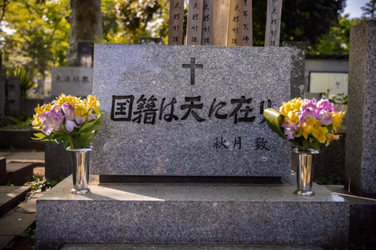 Headstone of Akitsuki Itaru with a cross and Japanese text, adorned with yellow and purple flowers in a cemetery.