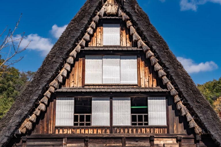 Traditional Japanese farmhouse with a steep thatched roof and white shoji windows against a clear blue sky.