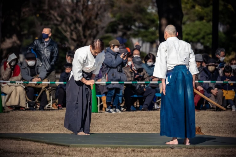 Aikido practitioners bow to each other during a Hama Rikyu aikido demonstration in a sunlit outdoor park.