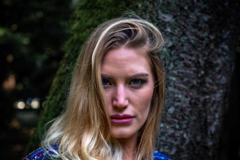 Close-up of a professional model with blonde hair leaning against a mossy tree and looking into the camera.