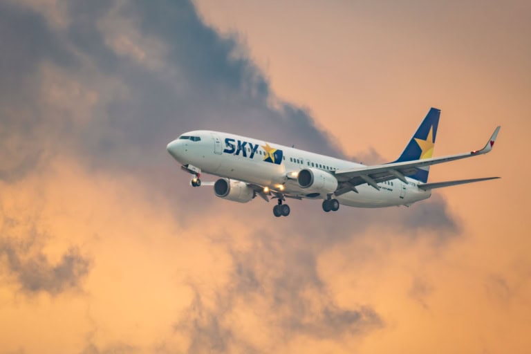 A Skymark Airlines plane descends at Jonanjima Seaside Park against a dramatic, golden sunset sky with dark clouds.