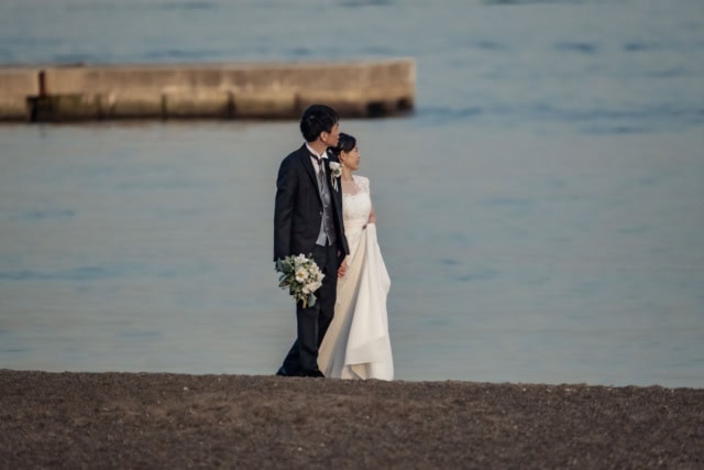 A bride and groom stand on a dark sand beach, looking out at calm water with a concrete pier in the distance.