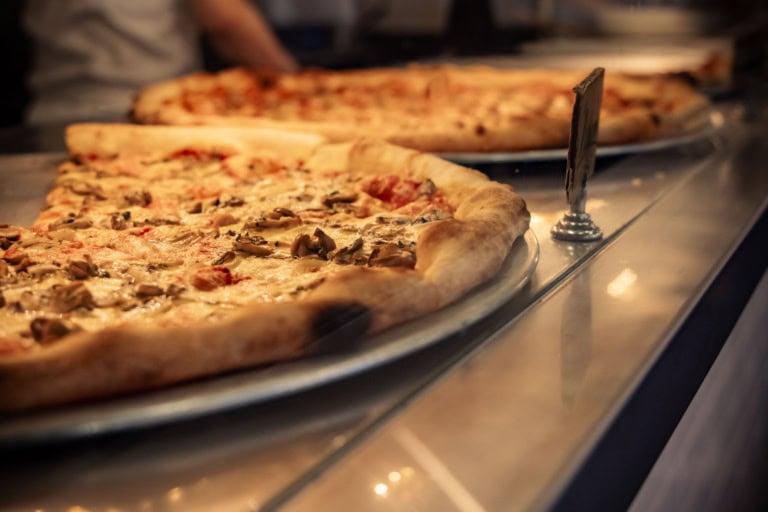 Close-up of a fresh mushroom pizza on a display counter, with another pizza blurred in the warm background.