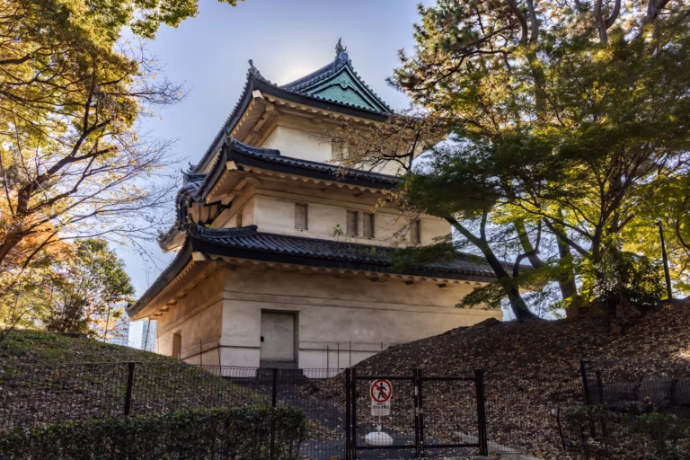A multi-tiered white Japanese watchtower with dark tiled roofs stands among autumn trees behind a black fence.