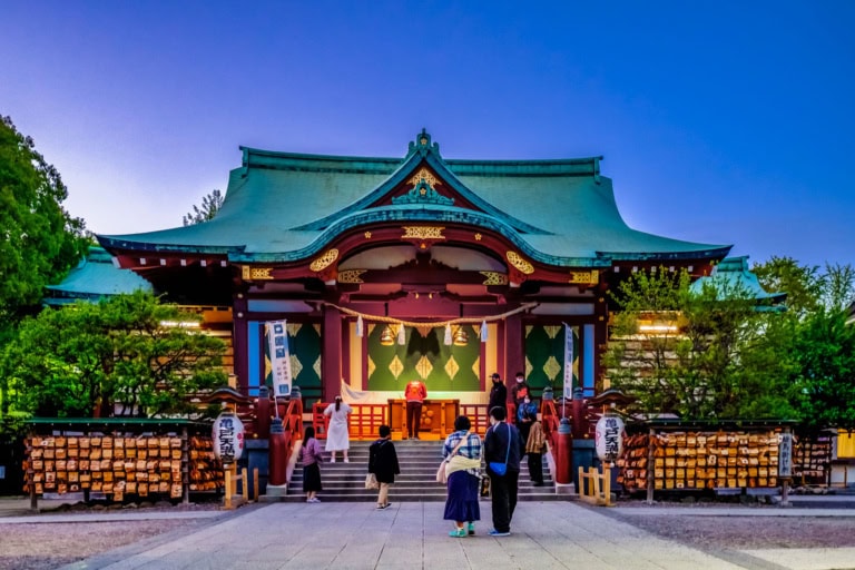 People walk toward a colorful Kameido Tenjin shrine with a green roof under a dark blue twilight sky.