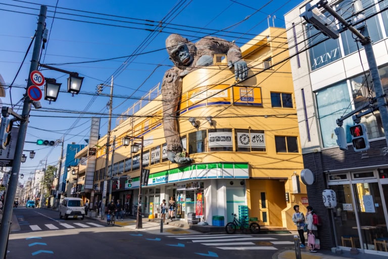 Giant King Kong statue sits above a yellow building with a FamilyMart on a sunny, urban street in Japan.