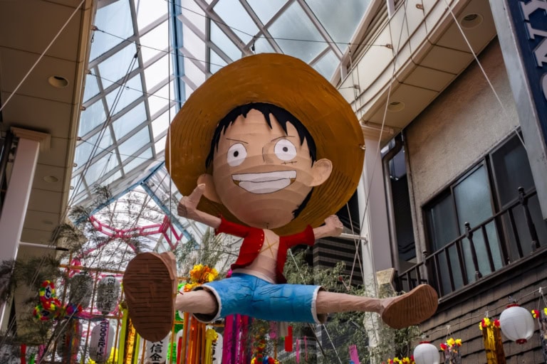 Large Luffy paper figure in a straw hat hanging at the Asagaya Tanabata Festival under a glass-roofed arcade.