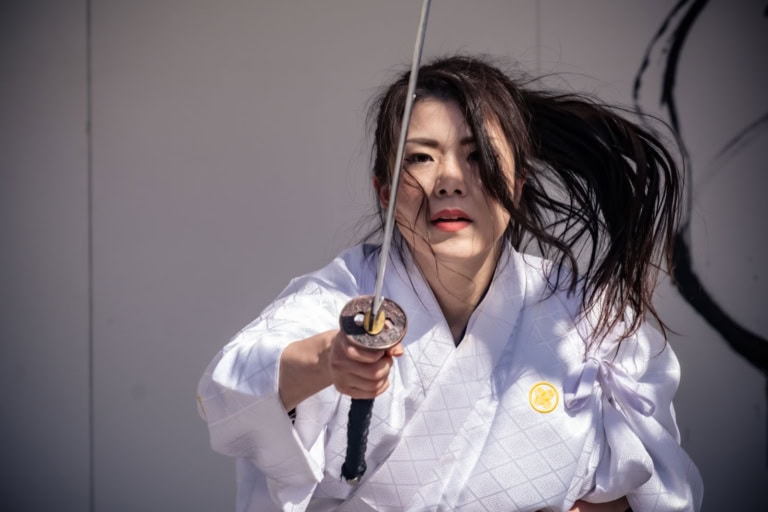 A woman in a white patterned kimono points a katana at the camera at Samurai Festival 2024, her hair windswept.