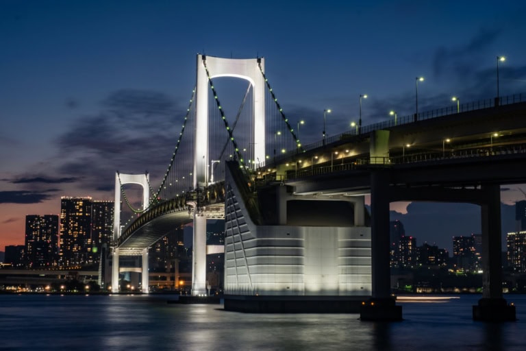 The illuminated Rainbow Bridge at night with white towers and green cable lights over the Tokyo Bay skyline.