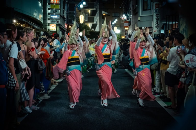 Dancers in pink kimonos and amigasa hats perform on a crowded street during the Shimokitazawa Awa Odori at night.