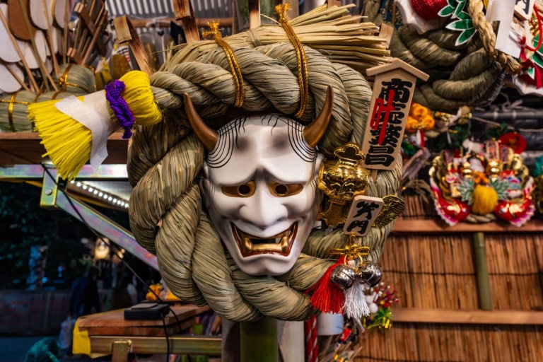 A white Hannya mask on a braided straw lucky rake with gold accents and charms at Hanazono Shrine’s San-no-Tori 2020.